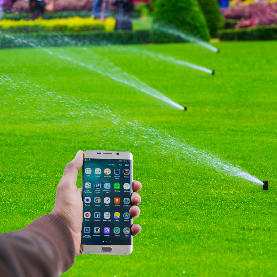 A hand holding a smartphone in front of a green lawn with multiple sprinklers spraying water, showing remote control of irrigation.